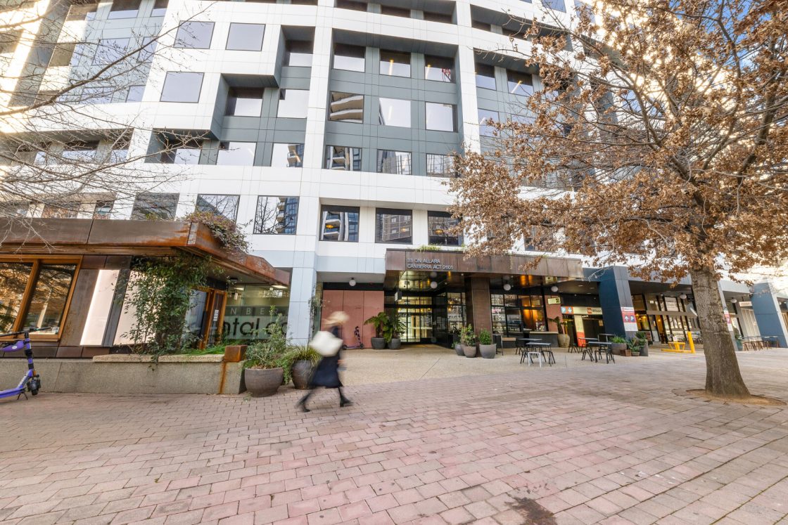 A person strolls across a plaza in front of a modern multi-story building housing various offices.