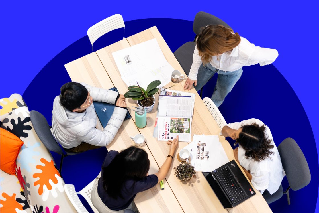 Four people are seated around a table with laptops, documents, and a plant, engaged in a discussion about web design. The background is a bold blue, and the chairs are white with a colorful patterned couch nearby.
