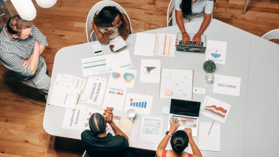 A group of people sit around a table with laptops and printed charts, discussing and analyzing data to create a marketing strategy.