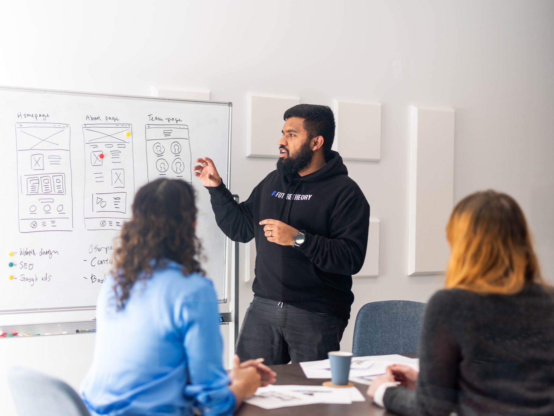 A man in a black hoodie explains diagrams on a whiteboard while two women seated at a table listen attentively.
