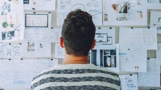 A person in a striped sweater examines a wall covered with papers, diagrams, and notes.